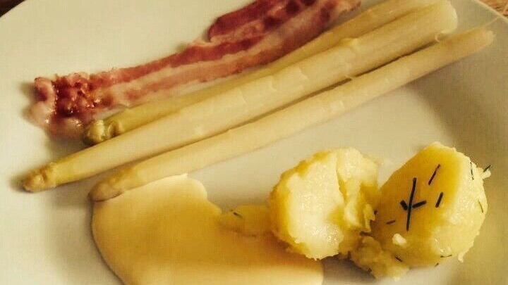 Ein Teller zeigt frisch gekochten Spargel mit Sauce und Kartoffeln, daneben liegen ein paar St&uuml;cke Speck, in einem rustikalen Umfeld.