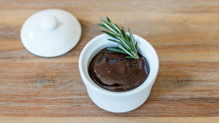 Eine kleine wei&szlig;e Sch&uuml;ssel mit dunklem Gelee, garniert mit frischem Rosmarin, steht auf einer Holzoberfl&auml;che.