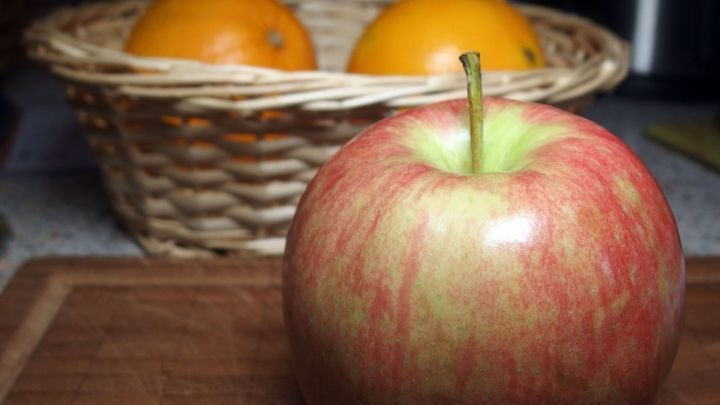 Ein roter Apfel liegt auf einem Holzbrett, w&auml;hrend im Hintergrund ein Korb mit Orangen steht, der frisches Obst zeigt.