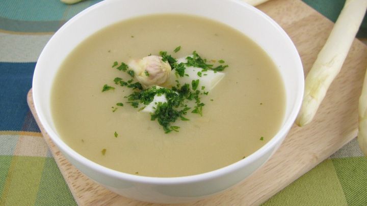 Eine Schale Spargelcremesuppe mit Petersilie garniert steht auf einem Holzbrett, umgeben von frischen Spargelstangen.
