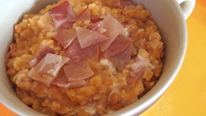 Eine sch&uuml;sselige Linsensuppe wird serviert, garniert mit Schinkenst&uuml;ckchen, auf einem bunten Untergrund.