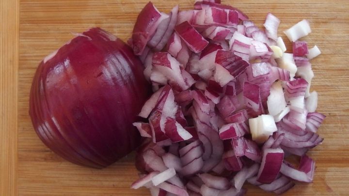 Ein roter Zwiebelw&uuml;rfel liegt neben fein gehackten Zwiebelst&uuml;cken auf einem Holzbrett in einer K&uuml;che.
