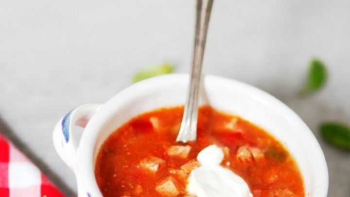 Eine Sch&uuml;ssel Soljanka mit einem L&ouml;ffel darin steht auf einem grauen Hintergrund, umgeben von einem karierten Tuch und frischem Basilikum.