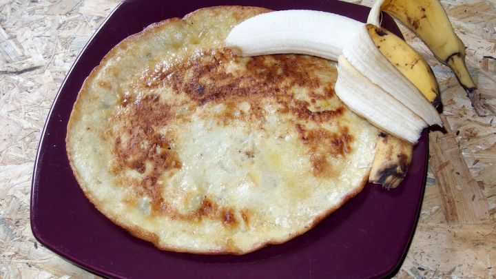 Auf einem lila Teller liegt ein goldbrauner Pfannkuchen, daneben ruhen eine ungesch&auml;lte und eine halbierte Banane, die verzehrbereit ist.