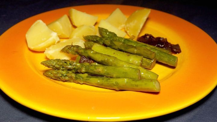 Gr&uuml;ner Spargel liegt auf einem bunten Teller neben Kartoffeln und einer dunklen Sauce, alles appetitlich angerichtet.