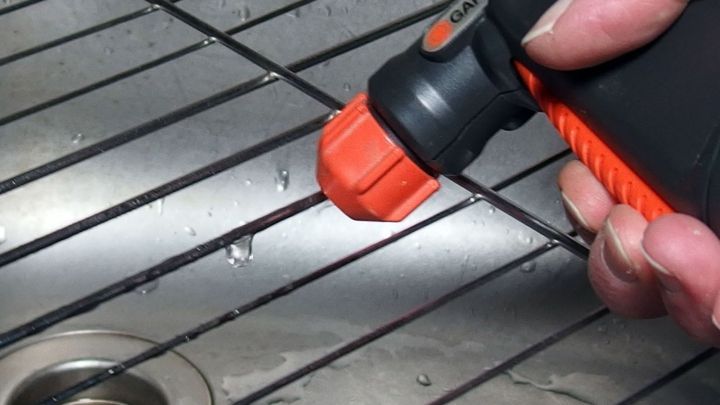 Ein Grillrost wird mit einem Spr&uuml;hger&auml;t gereinigt, w&auml;hrend Wasser in eine Ablauffl&auml;che darunter flie&szlig;t.