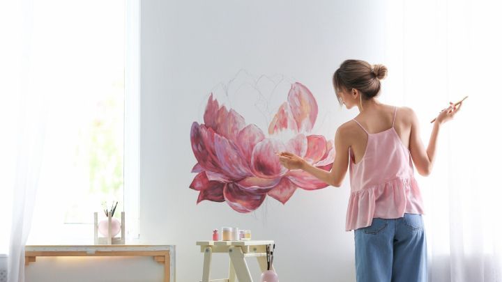 Eine Frau malt eine gro&szlig;e Blume an die Wand, w&auml;hrend sie auf einer Leiter in einem hellen Raum steht.