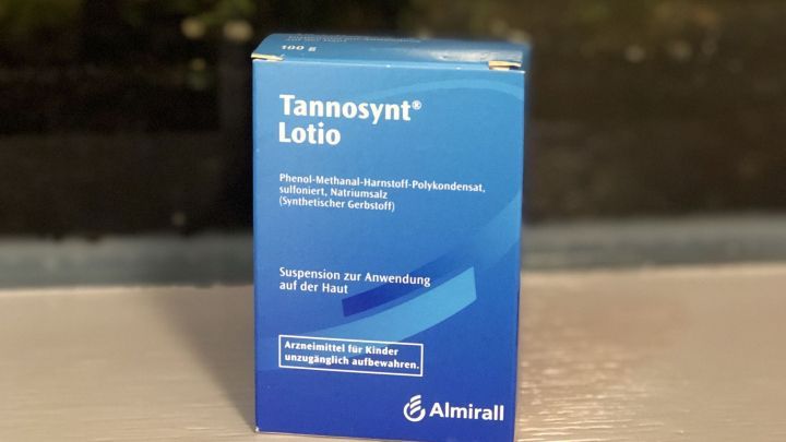 Eine blaue Verpackung von Tannosynt&reg; Lotion steht auf einem Tisch vor einer Pflanzenwand und wird als Behandlung gegen Dellwarzen genutzt.