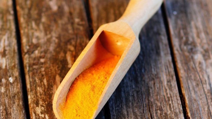 Eine h&ouml;lzerne Schaufel liegt auf einer rustikalen Holzoberfl&auml;che, gef&uuml;llt mit leuchtend gelbem Kurkuma-Pulver.