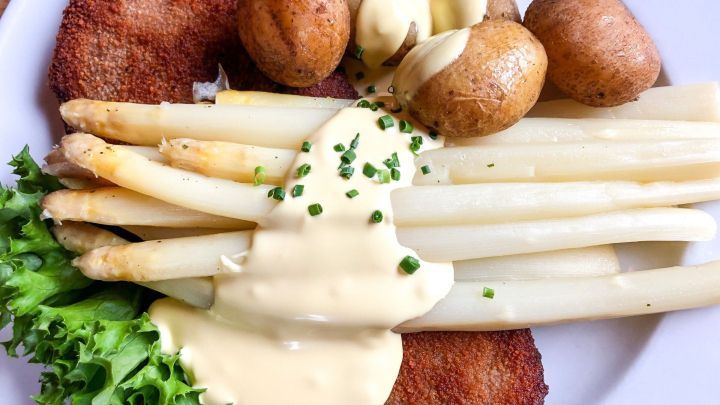 Zarte Spargelstangen liegen unter einer cremigen So&szlig;e, umgeben von Bratkartoffeln und einem Schnitzel auf einem Teller.