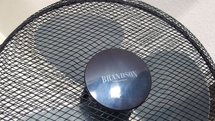 Ein Ventilator mit einem runden Kopf steht still da, umgeben von einem metallenen Schutzgitter in einem neutralen Raum.