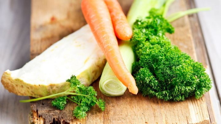 Frisches Suppengr&uuml;n liegt auf einem Holzbrett, w&auml;hrend Karotte, Lauch und Petersilie zur Eintopfzubereitung bereitstehen.