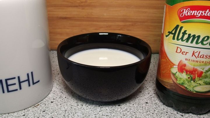 Eine schwarze Sch&uuml;ssel mit selbstgemachter saurer Sahne steht neben einer Flasche Essig und einem Beh&auml;lter f&uuml;r Mehl auf einer Arbeitsfl&auml;che.