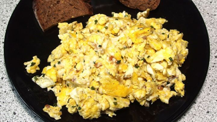 R&uuml;hreier liegen auf einem schwarzen Teller, w&auml;hrend zwei Scheiben Brot neben ihnen auf der Tischplatte liegen.