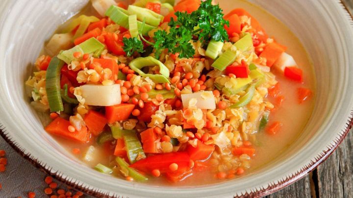 Ein bunter Gem&uuml;seeintopf mit roten Linsen liegt in einer Sch&uuml;ssel, umgeben von Holz und verstreuten Linsen.