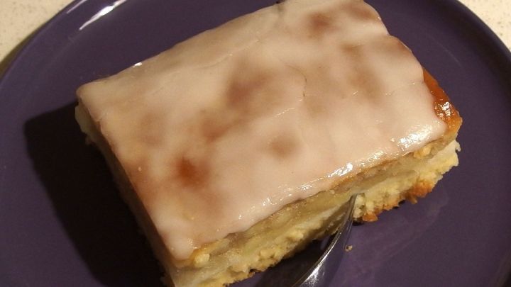 Ein St&uuml;ck Kuchen mit Zuckerguss aus Apfelsaft und Puderzucker liegt auf einem lila Teller in einer neutralen Umgebung.