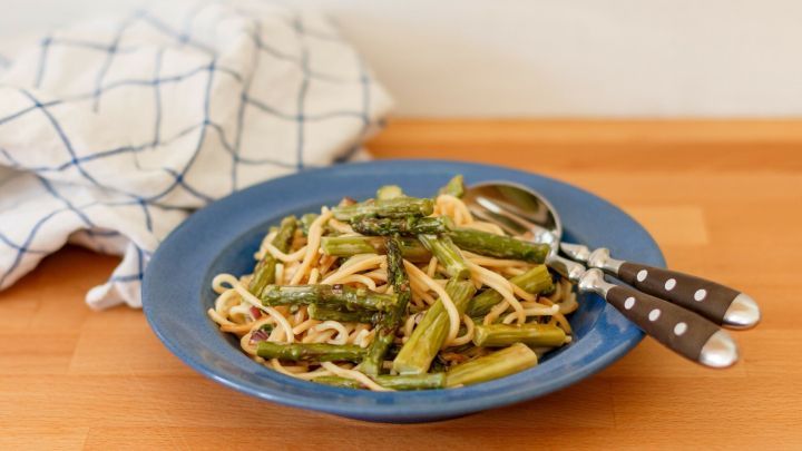 Ein Teller Nudeln mit frischem Spargel steht auf einem Holztisch, umgeben von einem karierten Geschirrtuch.
