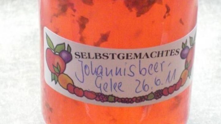 Ein Glas mit selbstgemachtem Johannisbeer-Prosecco-Gelee steht auf einer dunklen Fl&auml;che und zeigt leuchtend rote Farben.