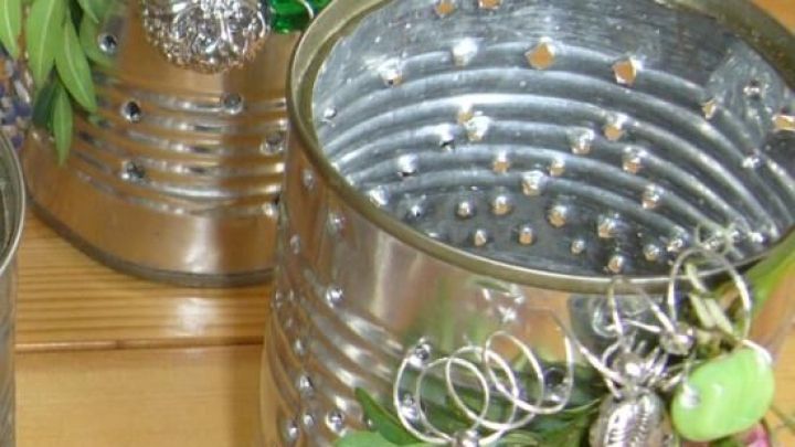 Zwei dekorierte Windlichter aus alten Konservendosen stehen auf einem Holztisch, umgeben von gr&uuml;nen Zweigen und Versch&ouml;nerungen.