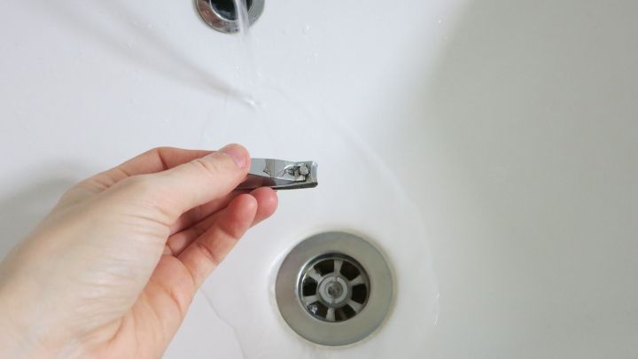 Eine Hand h&auml;lt einen Nagelknipser &uuml;ber einen Waschbeckenabfluss, w&auml;hrend Wasser hindurchflie&szlig;t.