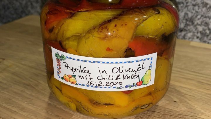 Ein Glas eingelegter Paprika steht auf einem Holzbrett, gef&uuml;llt mit bunten Paprikast&uuml;cken und eingelegt in &Ouml;l.