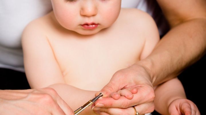 Kleinkind sitzt auf dem Scho&szlig; eines Erwachsenen, w&auml;hrend eine Hand sanft die N&auml;gel des Kindes mit einer Schere schneidet.