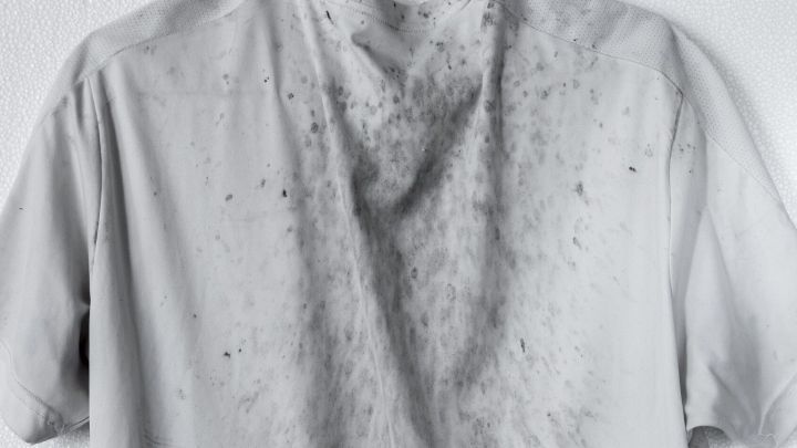 Ein verschmutztes, graues T-Shirt mit Schmier- und &Ouml;lflecken h&auml;ngt an einer Wand.