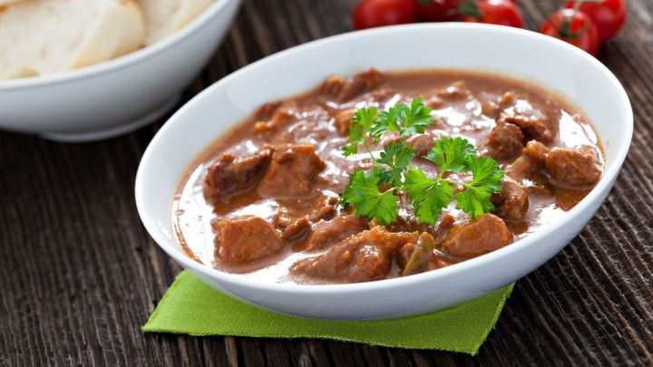 Ein schmackhaftes Pferdegulasch mit Kabeln und frischer Petersilie in einer wei&szlig;en Sch&uuml;ssel, umgeben von Brot und Tomaten.