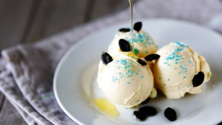 Drei Kugeln Vanilleeis werden mit K&uuml;rbiskernen und K&uuml;rbis&ouml;l auf einem wei&szlig;en Teller verziert, umgeben von einem grauen Tuch.