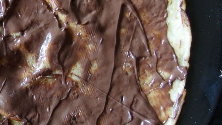 Auf einer Pfanne liegt ein goldbraun gebratener Pfannkuchen, der gro&szlig;z&uuml;gig mit Schokoso&szlig;e &uuml;berzogen ist.