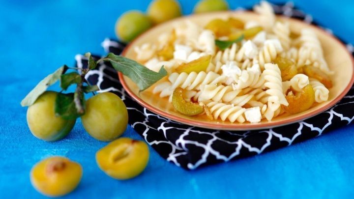 Auf einem Holzbrett liegen s&uuml;&szlig;e Nudeln mit Mirabellen und Ziegenfrischk&auml;se, umgeben von frischen Mirabellen und einem bunten Hintergrund.