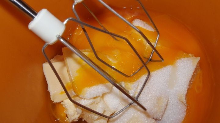 Ein Mixer mit R&uuml;hrbesen steht &uuml;ber einer Sch&uuml;ssel mit Zucker und Eiern, w&auml;hrend die Zutaten f&uuml;r den Mohnkuchen vorbereitet werden.