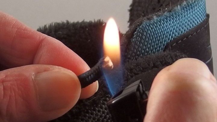 Eine Hand h&auml;lt ausgefranste Schn&uuml;rsenkel &uuml;ber einer Flamme, um sie zu reparieren. Der Hintergrund ist unscharf und neutral.