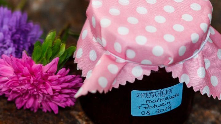 Ein Glas Zwetschgen-Rotweinmarmelade mit einem gepolsterten, pinken Deckel steht auf einem rustikalen Untergrund, umgeben von bunten Blumen.