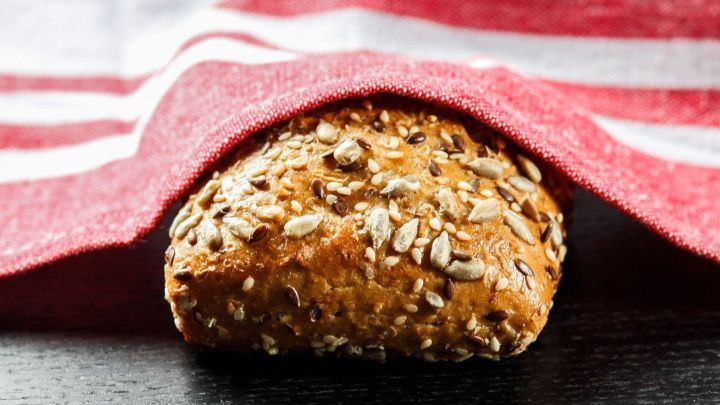 Ein goldbraunes Br&ouml;tchen mit Saaten liegt unter einem rot-wei&szlig;en Geschirrtuch auf einem dunklen Tisch.