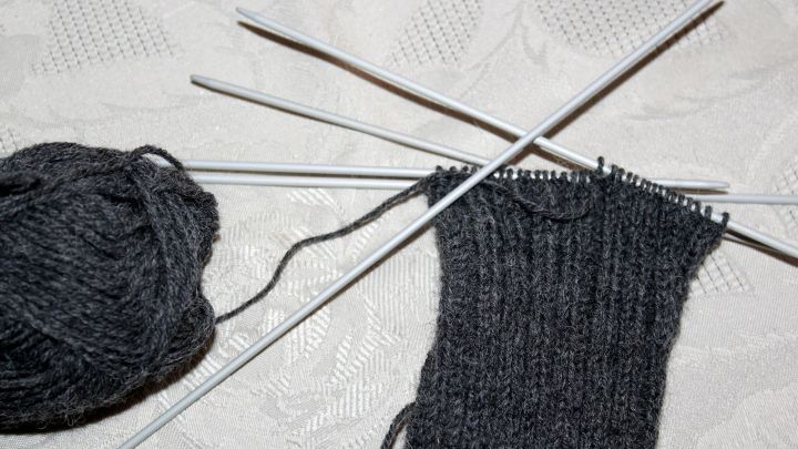 Dunkles Garn und Stricknadeln liegen auf einem Tisch, w&auml;hrend ein &Auml;rmel in Ribbelmuster langsam entsteht.