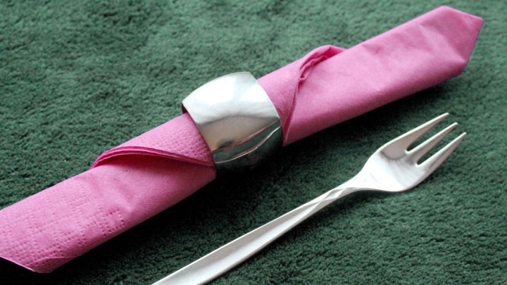 Eine silberne Serviettenring liegt um eine pinke Serviette, w&auml;hrend eine Gabel auf einem gr&uuml;nen Tisch liegt.