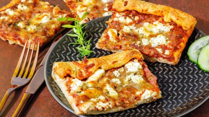 Ein Teller mit viereckigen Pizzast&uuml;cken, die k&ouml;stlich belegt sind, steht auf einem rustikalen Tisch neben einer Gabel und Gurkenscheiben.