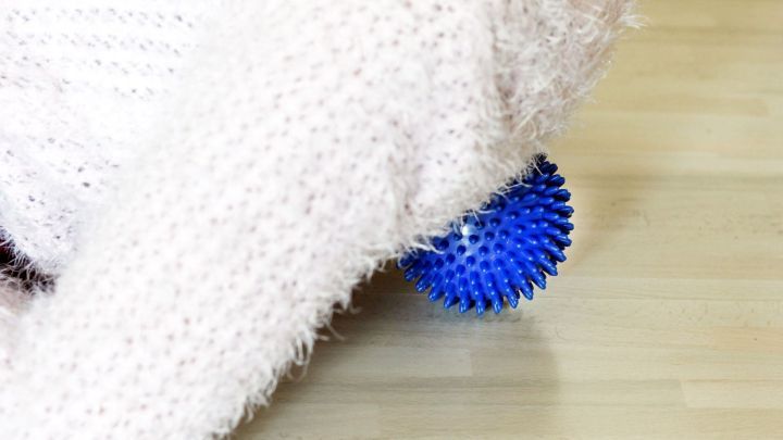 Ein blauer Massageball liegt auf einem Holztisch, w&auml;hrend eine Hand mit einem flauschigen Pullover ihn sanft dr&uuml;ckt, um Verspannungen zu l&ouml;sen.