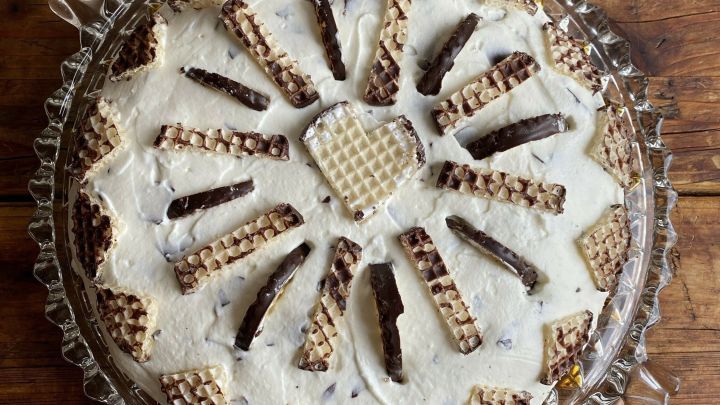 Eine Schokokuss-Torte ist auf einem Glasplattenrand platziert, dekoriert mit Waffelst&uuml;cken und Sahne, vor einem Holztisch.