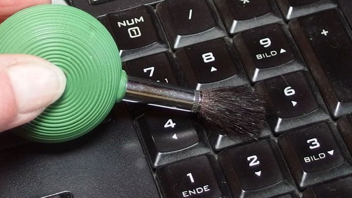 Ein kleiner, gr&uuml;ner Blasebalg mit Pinsel reinigt ein Keyboard, w&auml;hrend eine Hand das Ger&auml;t f&uuml;hrt.