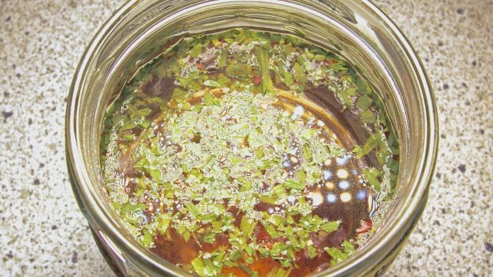 Ein Glas mit Essig und Kr&auml;utern steht auf einer Arbeitsfl&auml;che, w&auml;hrend die Zutaten zum Verfeinern des Essigs vermischt sind.