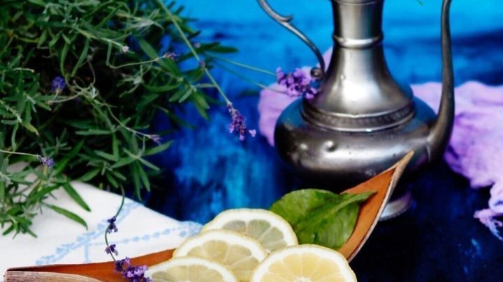 Eine silberne Kanne steht neben frischen Zitronenscheiben und Kr&auml;utern auf einem wei&szlig;en Tuch, umgeben von einer blauen Kulisse.