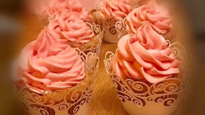 Kleine Muffins mit rosa Sahnehaube stehen auf einem Holztisch, im Hintergrund unscharf gefiltert, festlich pr&auml;sentiert.