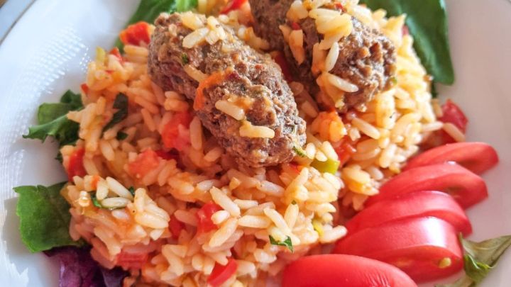 Saftige Hackfleischb&auml;llchen liegen auf einem bunten Reisgericht, umgeben von frischen Tomatenscheiben und Blattsalat.