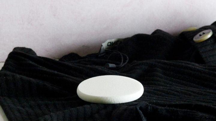 Ein runder, wei&szlig;er Fleckenentferner liegt auf einem schwarzen Kleidungsst&uuml;ck auf einem Tisch vor einer hellen Wand.