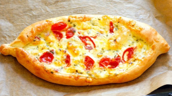 Ein knuspriger, ovaler Teig mit Schmand und Tomatenfr&uuml;chten liegt auf einem Backblech, umgeben von Backpapier.