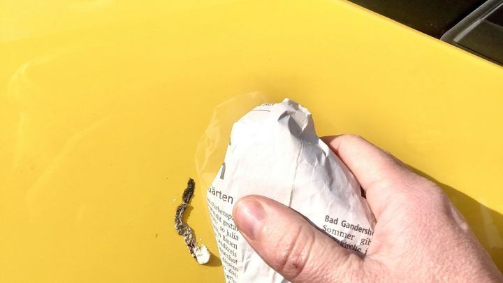 Ein Hand mit Zeitungspapier entfernt Vogelkacke von einem gelben Auto in einem sonnigen Umfeld.
