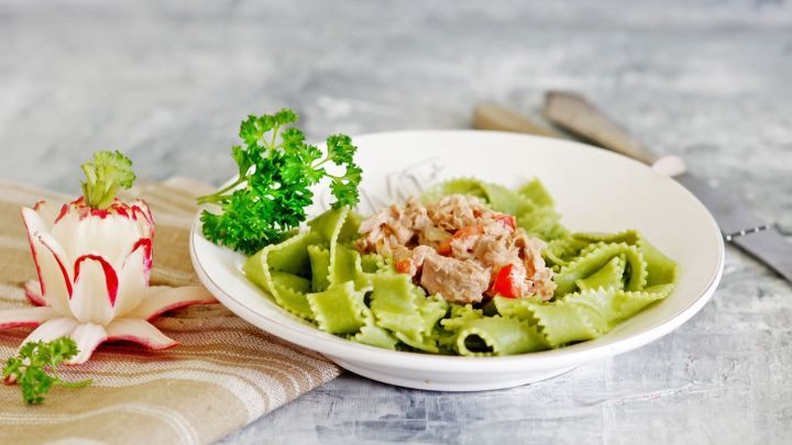 Gr&uuml;ne Bandnudeln liegen in einer wei&szlig;en Sch&uuml;ssel und sind mit einer cremigen Thunfischsauce und frischem Gem&uuml;se garniert.