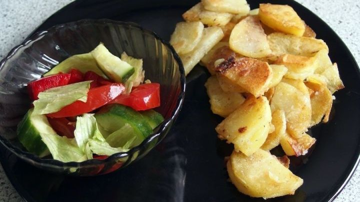 Auf einem schwarzen Teller liegen goldbraun gebratene Kartoffeln neben einer Sch&uuml;ssel mit frischem Salat aus verschiedenen Gem&uuml;sesorten.